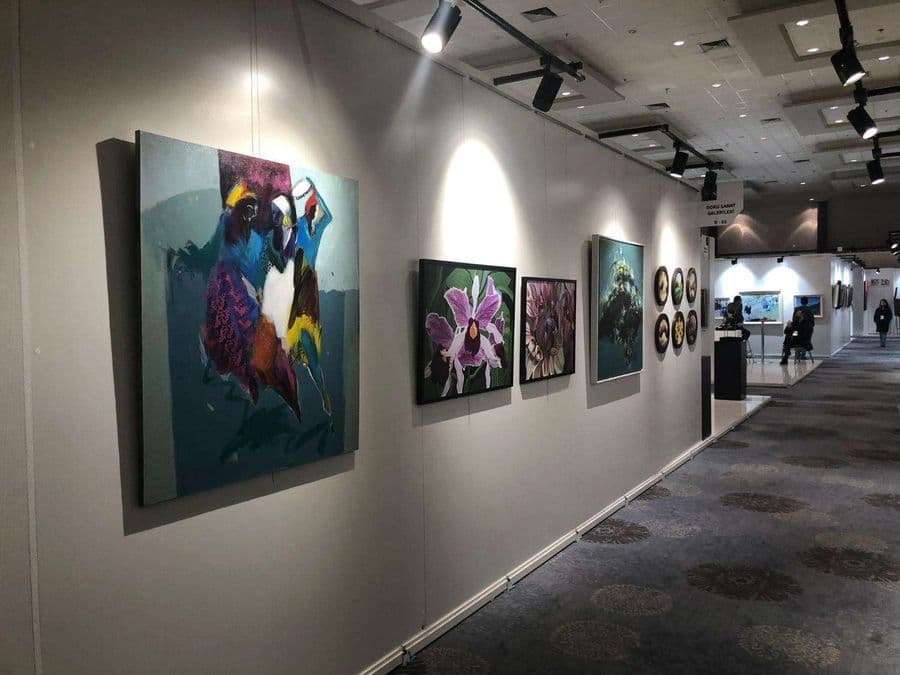 Desenli halı kaplı bir fuar koridorunun sol duvarında, spot ışıklarıyla aydınlatılan bir dizi sanat eseri sergileniyor. Eserler arasında soyut ve çiçek temalı tablolar bulunuyor.