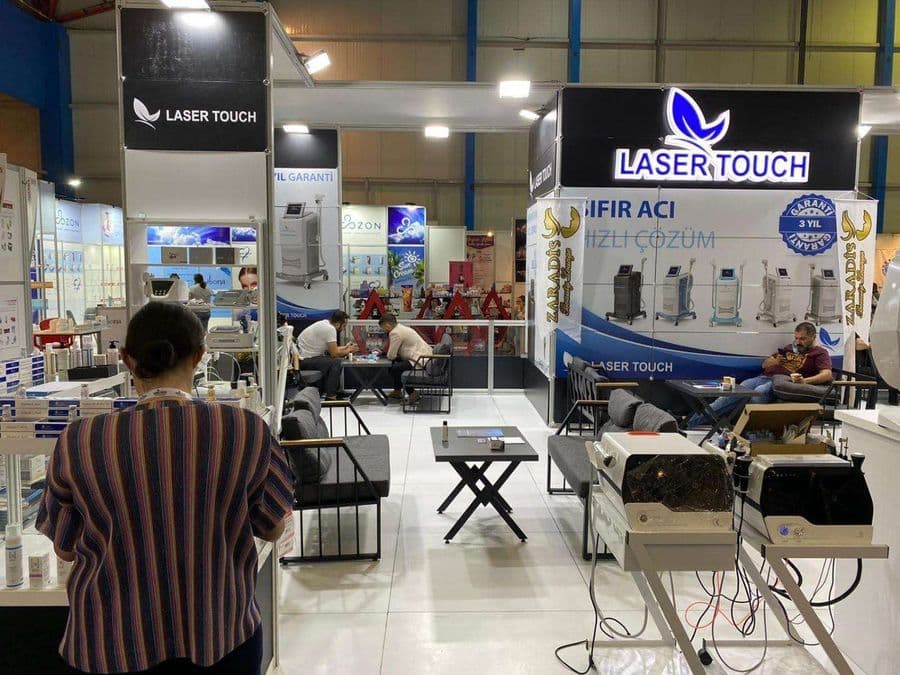 "Laser Touch" markasına ait, estetik ve lazer cihazlarının sergilendiği, kapalı bir ticaret fuarı standı. Standın sağ tarafında "2500W Yeni Nesil IPL" yazılı büyük bir tabela ve bir cihaz sergileniyor. Beyaz raflarda kozmetik ürünler, stant zemininde ise "ICEBERG" yazılı bir cihaz da dahil olmak üzere estetik cihazlar yer alıyor.
