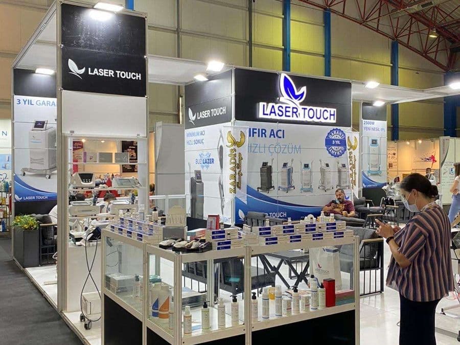 "Laser Touch" markasına ait, estetik ve lazer cihazlarının sergilendiği, kapalı bir ticaret fuarı standı. Standın sağ tarafında "2500W Yeni Nesil IPL" yazılı büyük bir tabela ve bir cihaz sergileniyor. Beyaz raflarda kozmetik ürünler, stant zemininde ise "ICEBERG" yazılı bir cihaz da dahil olmak üzere estetik cihazlar yer alıyor.