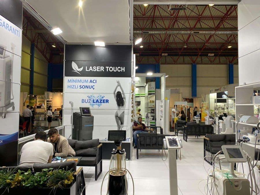 "Laser Touch" markasına ait, estetik ve lazer cihazlarının sergilendiği, kapalı bir ticaret fuarı standı. Standın sağ tarafında "2500W Yeni Nesil IPL" yazılı büyük bir tabela ve bir cihaz sergileniyor. Beyaz raflarda kozmetik ürünler, stant zemininde ise "ICEBERG" yazılı bir cihaz da dahil olmak üzere estetik cihazlar yer alıyor.