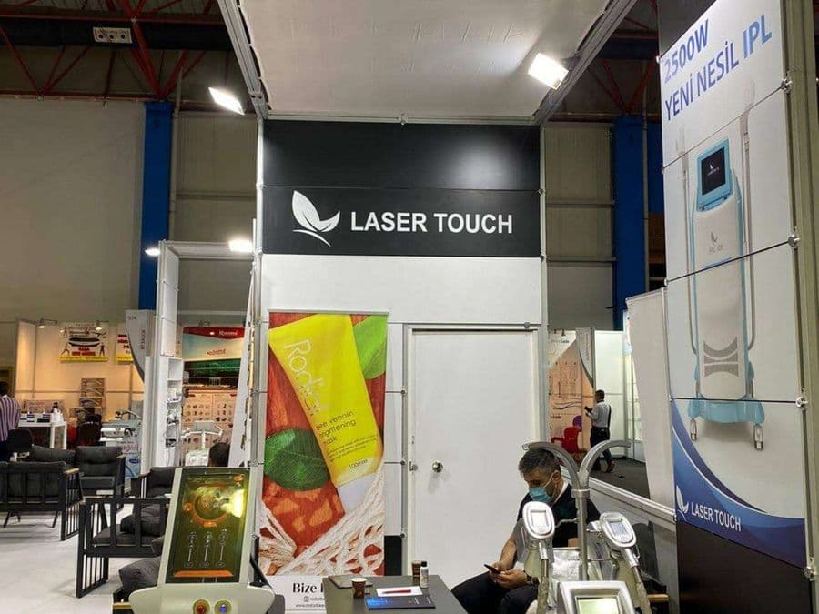 "Laser Touch" markasına ait, estetik ve lazer cihazlarının sergilendiği, kapalı bir ticaret fuarı standı. Standın sağ tarafında "2500W Yeni Nesil IPL" yazılı büyük bir tabela ve bir cihaz sergileniyor. Beyaz raflarda kozmetik ürünler, stant zemininde ise "ICEBERG" yazılı bir cihaz da dahil olmak üzere estetik cihazlar yer alıyor.