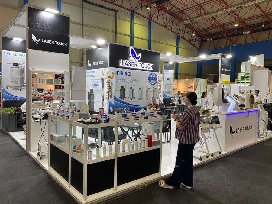 "Laser Touch" markasına ait, estetik ve lazer cihazlarının sergilendiği, kapalı bir ticaret fuarı standı. Standın sağ tarafında "2500W Yeni Nesil IPL" yazılı büyük bir tabela ve bir cihaz sergileniyor. Beyaz raflarda kozmetik ürünler, stant zemininde ise "ICEBERG" yazılı bir cihaz da dahil olmak üzere estetik cihazlar yer alıyor.