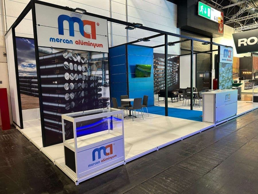 Kapalı bir fuar alanında kurulmuş, "mG Merçan Alüminyum" markasına ait profesyonel bir ticaret fuarı standı. Standın ön yüzünde paletlenmiş alüminyum külçelerinin büyük bir görseli yer alıyor. Stand, bir karşılama bankosu, oturma/toplantı alanı ve siyah metal çerçeveli modern bir tasarıma sahiptir. Zemin koyu renkli beton ve stant alanı beyaz ve açık mavi halı ile kaplıdır.
