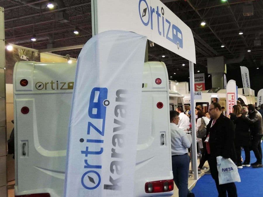 Bir karavan fuarı veya ticaret fuarı standında sergilenen, beyaz renkli, "Ortiz" markalı bir çekme karavan (römork karavan). Mavi halı kaplı alanda karavanın etrafında yürüyen ve inceleyen ziyaretçiler bulunuyor.