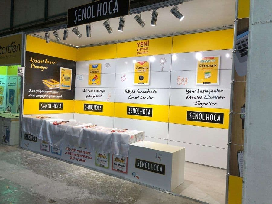 "Şenol Hoca" eğitim markasının bir fuardaki kitap satış standı. Sarı ve beyaz renklerin hakim olduğu stantta, matematik kitapları sergileniyor ve duvarlarda öğrencilere yönelik sloganlar yer alıyor.