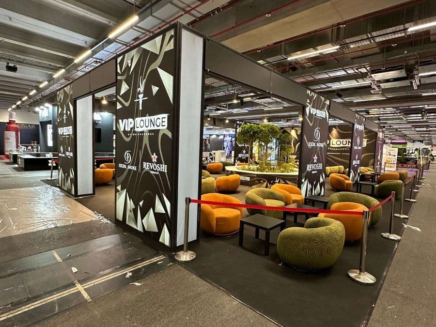 Bir fuar veya kongre merkezinde, kırmızı şeritlerle ayrılmış "VIP Lounge" dinlenme bölümü. Alanda, turuncu ve yeşil renklerde çok sayıda yuvarlak hatlı koltuk ve küçük sehpalar bulunuyor.