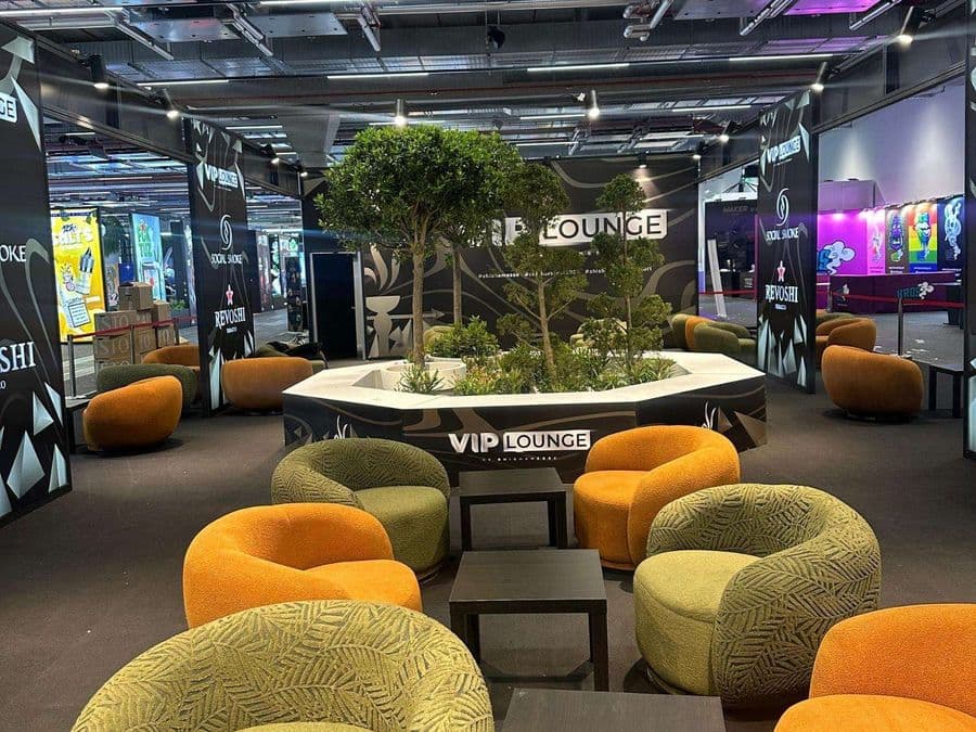 Bir fuar veya kongre merkezinde, kırmızı şeritlerle ayrılmış "VIP Lounge" dinlenme bölümü. Alanda, turuncu ve yeşil renklerde çok sayıda yuvarlak hatlı koltuk ve küçük sehpalar bulunuyor.