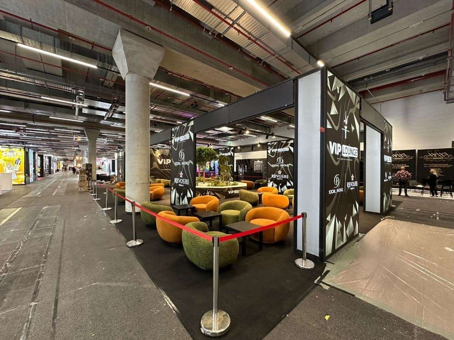 Bir fuar veya kongre merkezinde, kırmızı şeritlerle ayrılmış "VIP Lounge" dinlenme bölümü. Alanda, turuncu ve yeşil renklerde çok sayıda yuvarlak hatlı koltuk ve küçük sehpalar bulunuyor.