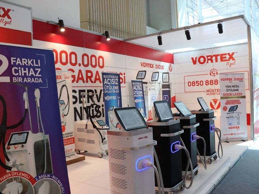 "Vortex Diode Lazer" markasının medikal estetik cihazlarını sergilediği bir fuar standı. Kırmızı ve beyaz temalı stantta, çok sayıda lazer cihazı yan yana dizilerek tanıtılıyor.
