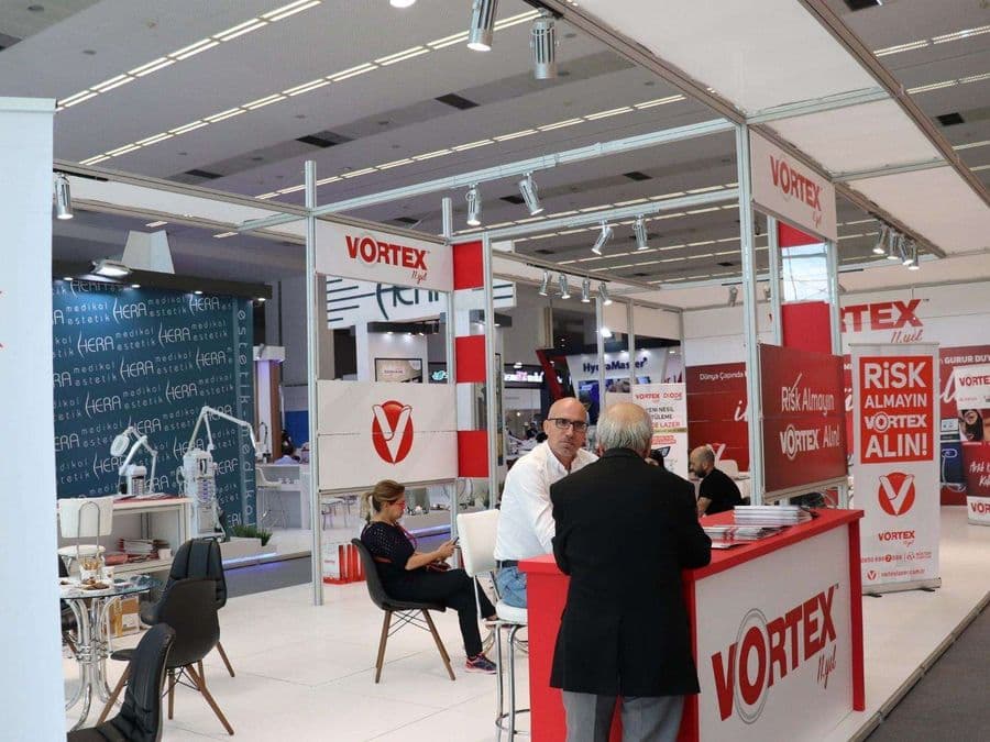 Endüstriyel ürünler fuarında, "VORTEX" markasının modern standı. Işıklı büyük bir logoya sahip standın içi, ziyaretçiler için çok sayıda kanepe ve koltukla bir bekleme alanı gibi tasarlanmış.