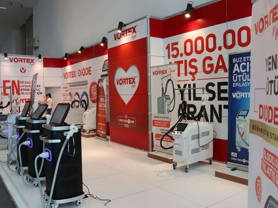 "Vortex Diode Lazer" markasının medikal estetik cihazlarını sergilediği bir fuar standı. Kırmızı ve beyaz temalı stantta, çok sayıda lazer cihazı yan yana dizilerek tanıtılıyor.