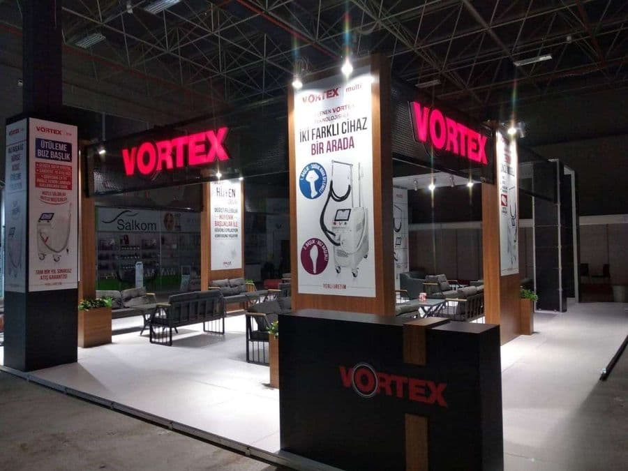 Endüstriyel ürünler fuarında, "VORTEX" markasının modern standı. Işıklı büyük bir logoya sahip standın içi, ziyaretçiler için çok sayıda kanepe ve koltukla bir bekleme alanı gibi tasarlanmış.
